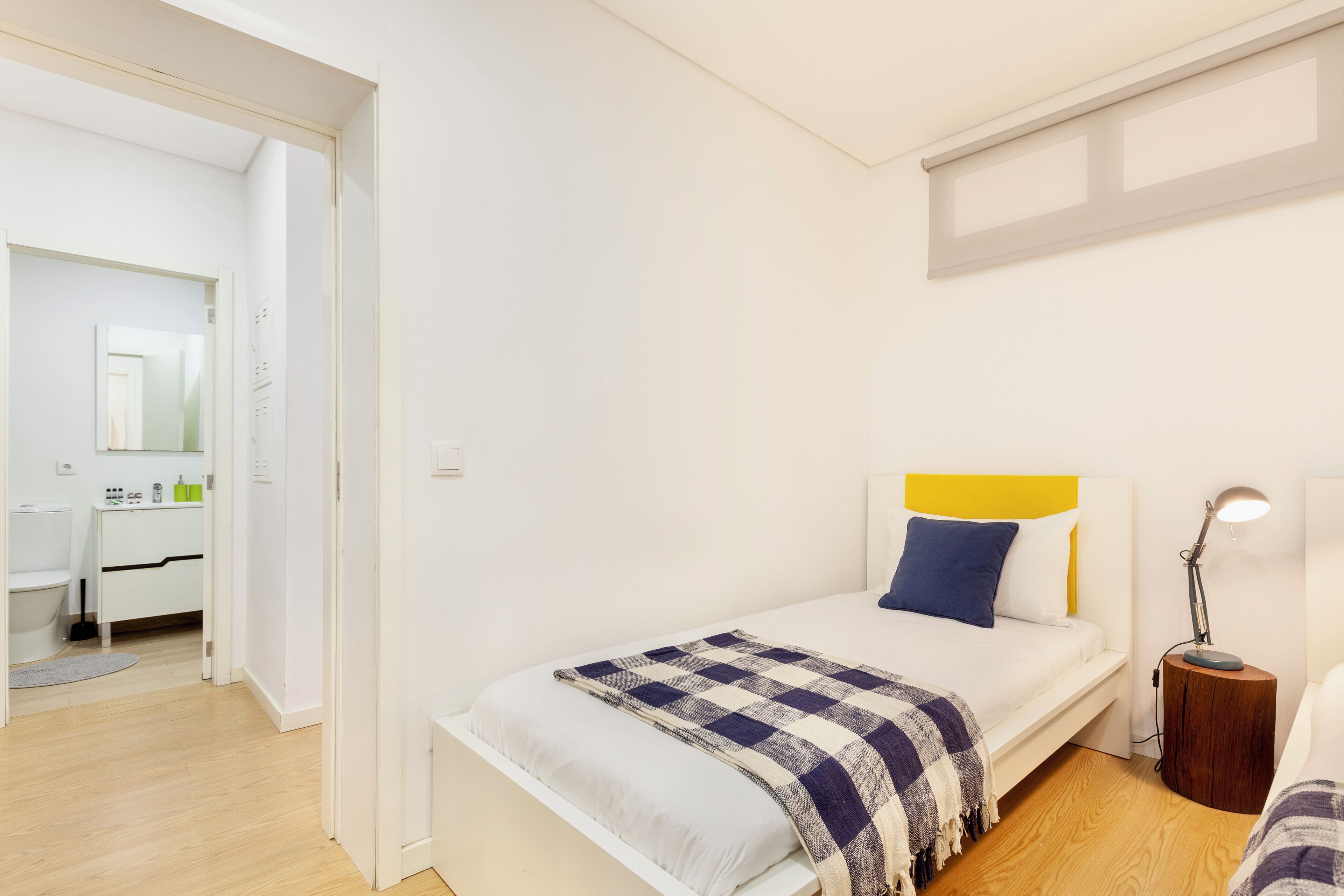 A bedroom with a bed, bedside table, and a lamp. There is a bathroom visible in the background.