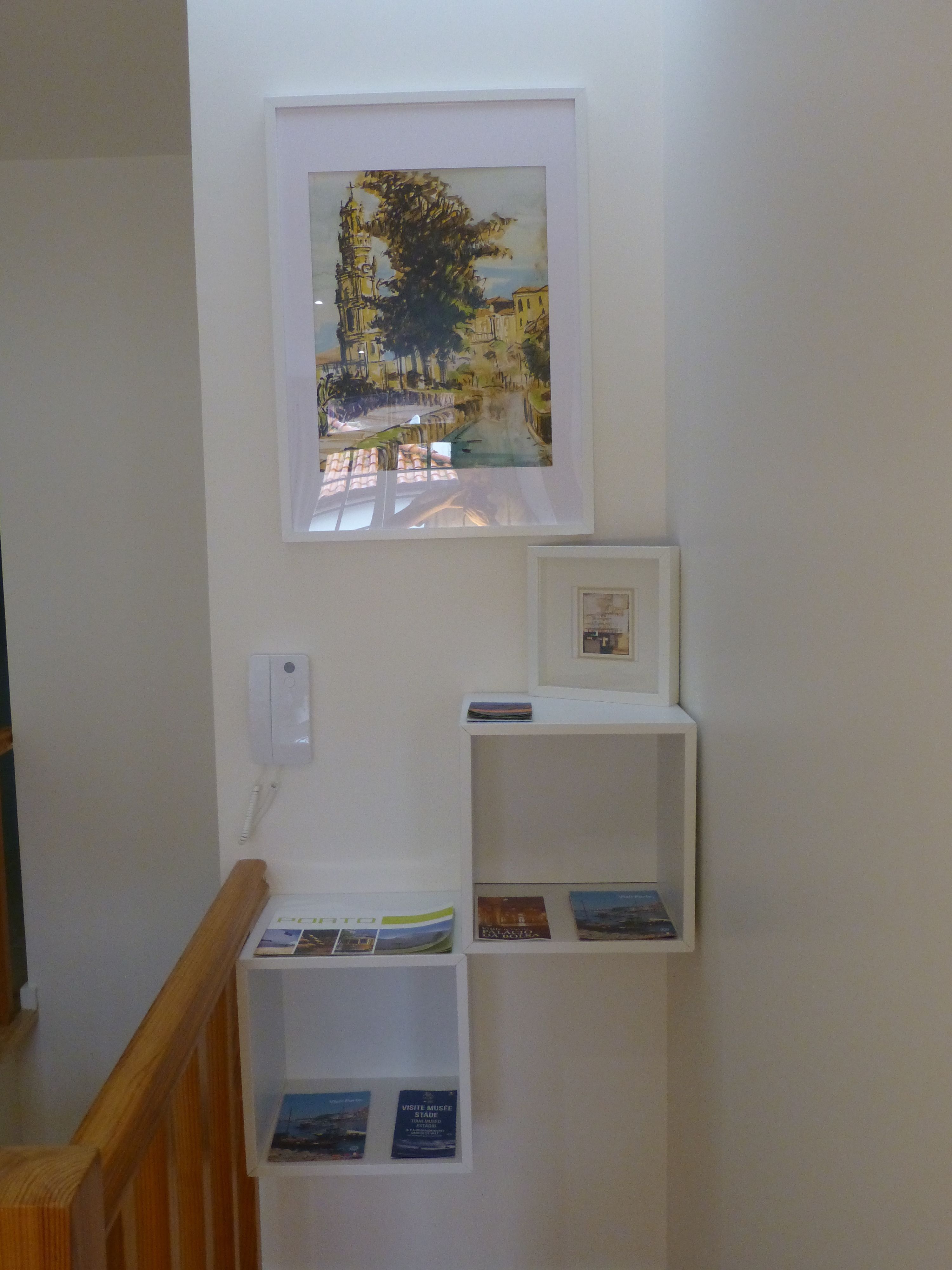 A staircase with a framed painting of a street scene, a white wall-mounted phone, and two white shelves with brochures.