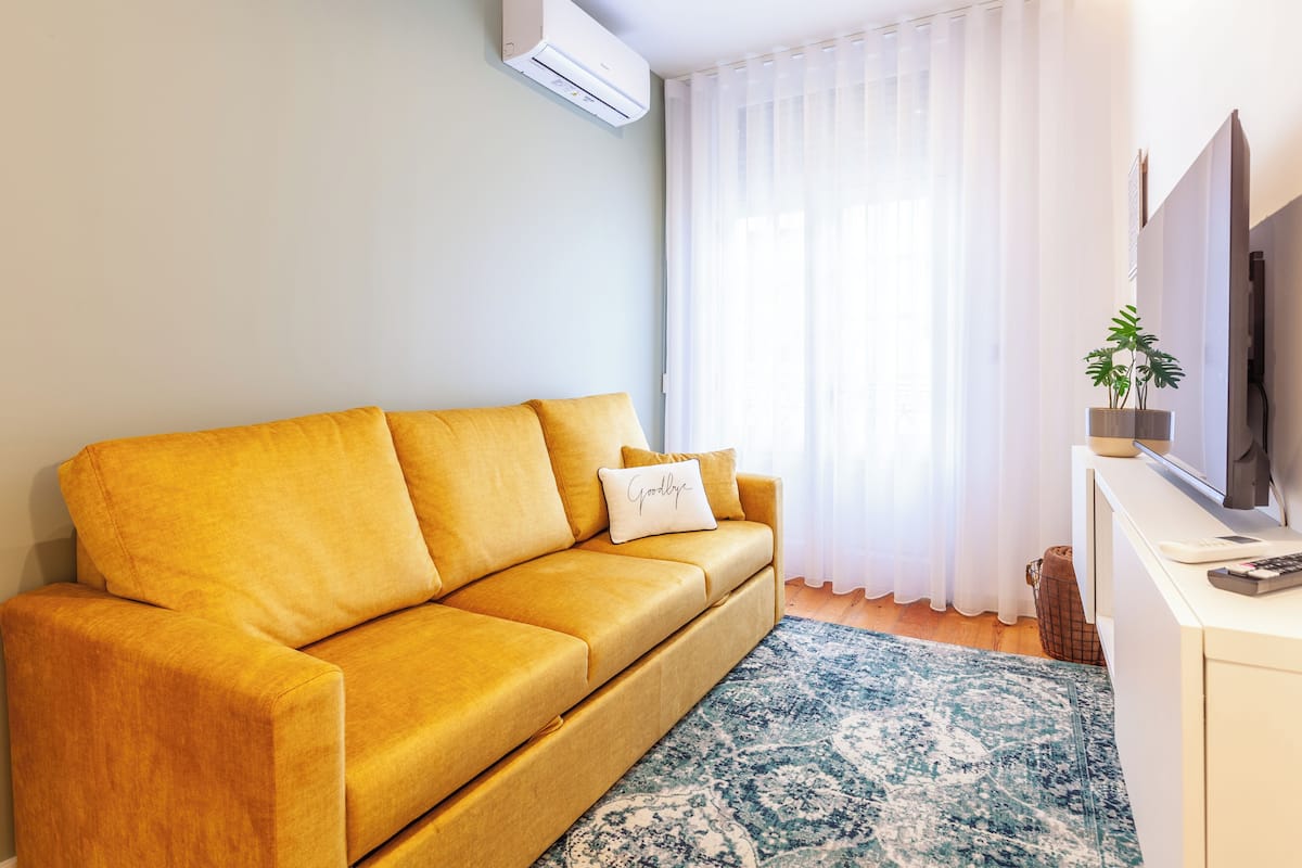 A yellow sofa with a 'Goodbye' pillow, a TV on a white stand, and a potted plant.