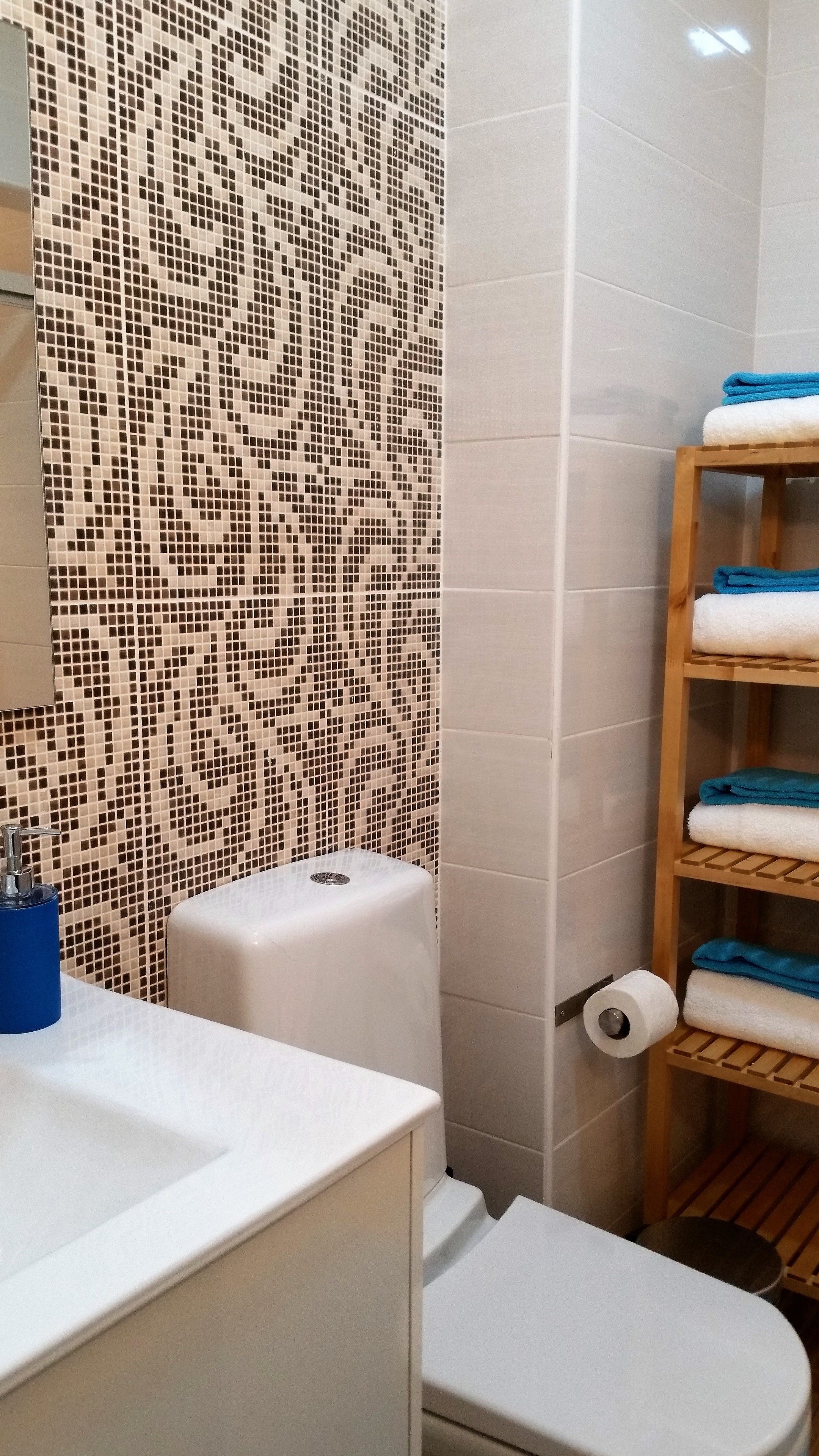 A bathroom with a mosaic tile wall, a toilet, a sink, and a shelf with towels.