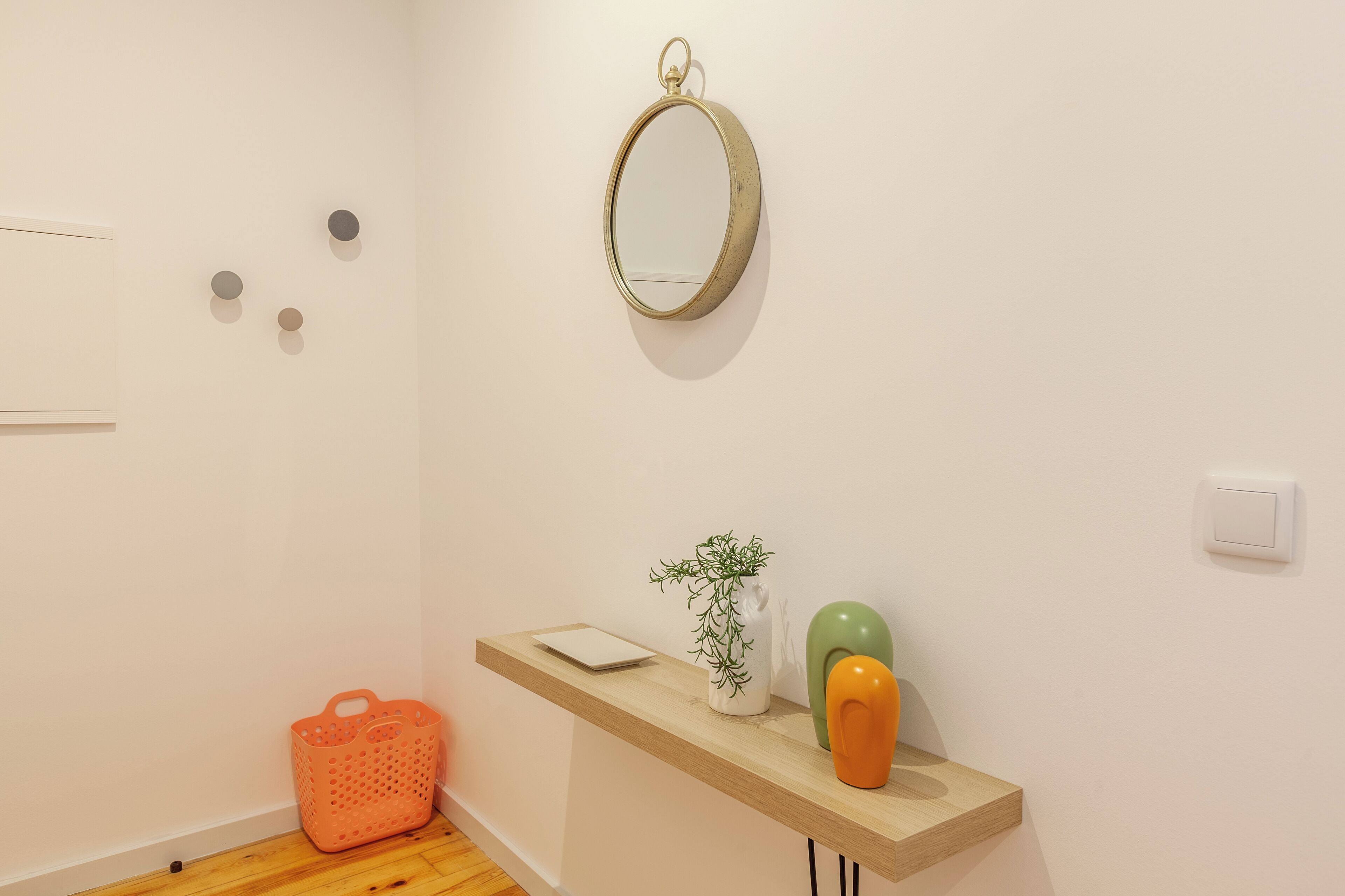 Un couloir avec un plancher en bois, un mur blanc, un miroir, une étagère avec une plante, un vase et un panier.