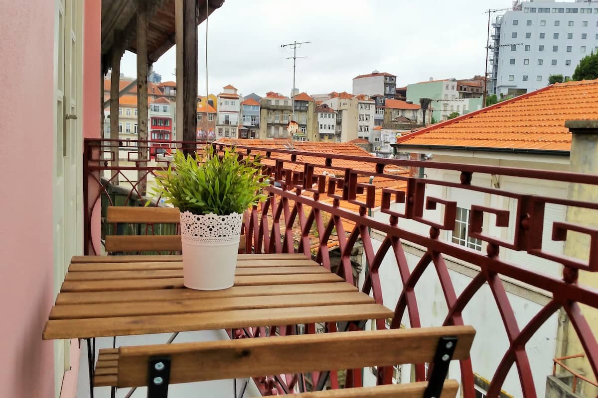 A balcony with a wooden table and a potted plant, overlooking a cityscape with terracotta-roofed buildings.
