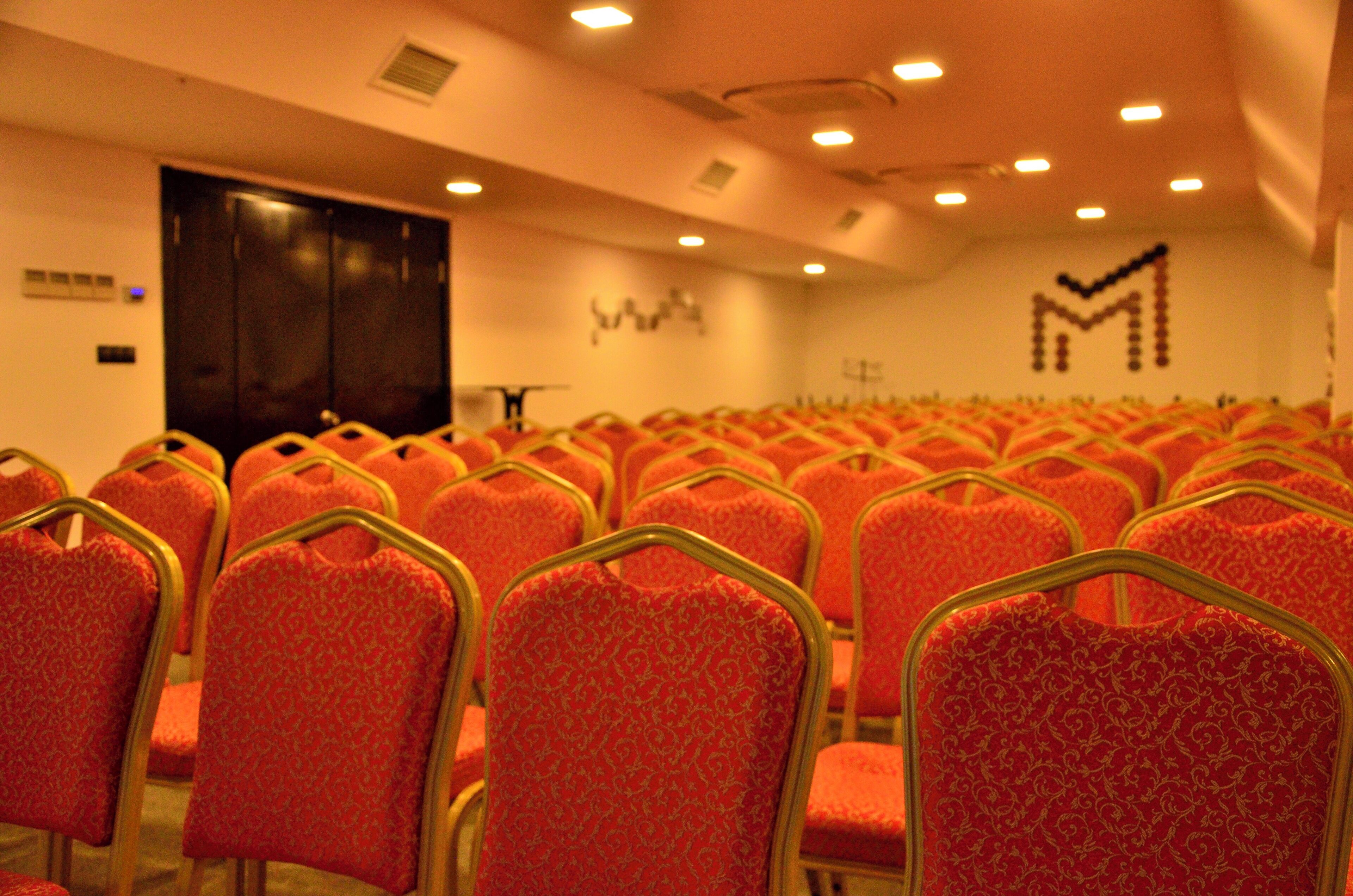 Rows of red chairs with gold frames in a conference room with a dark door and a wall with a geometric pattern.