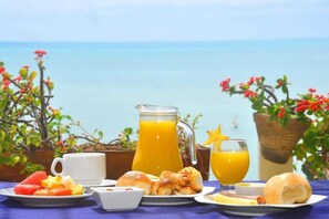 Un plateau de petit-déjeuner comprenant une carafe de jus d’orange, un verre de jus d’orange, des croissants et une assiette de fruits, le tout disposé sur une table offrant une vue sur la mer.