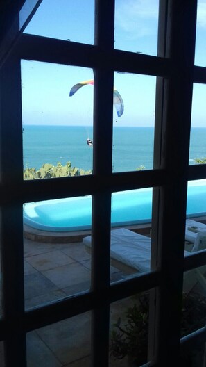Une vue à travers une fenêtre sur une piscine, avec une personne pratiquant le parapente au loin.