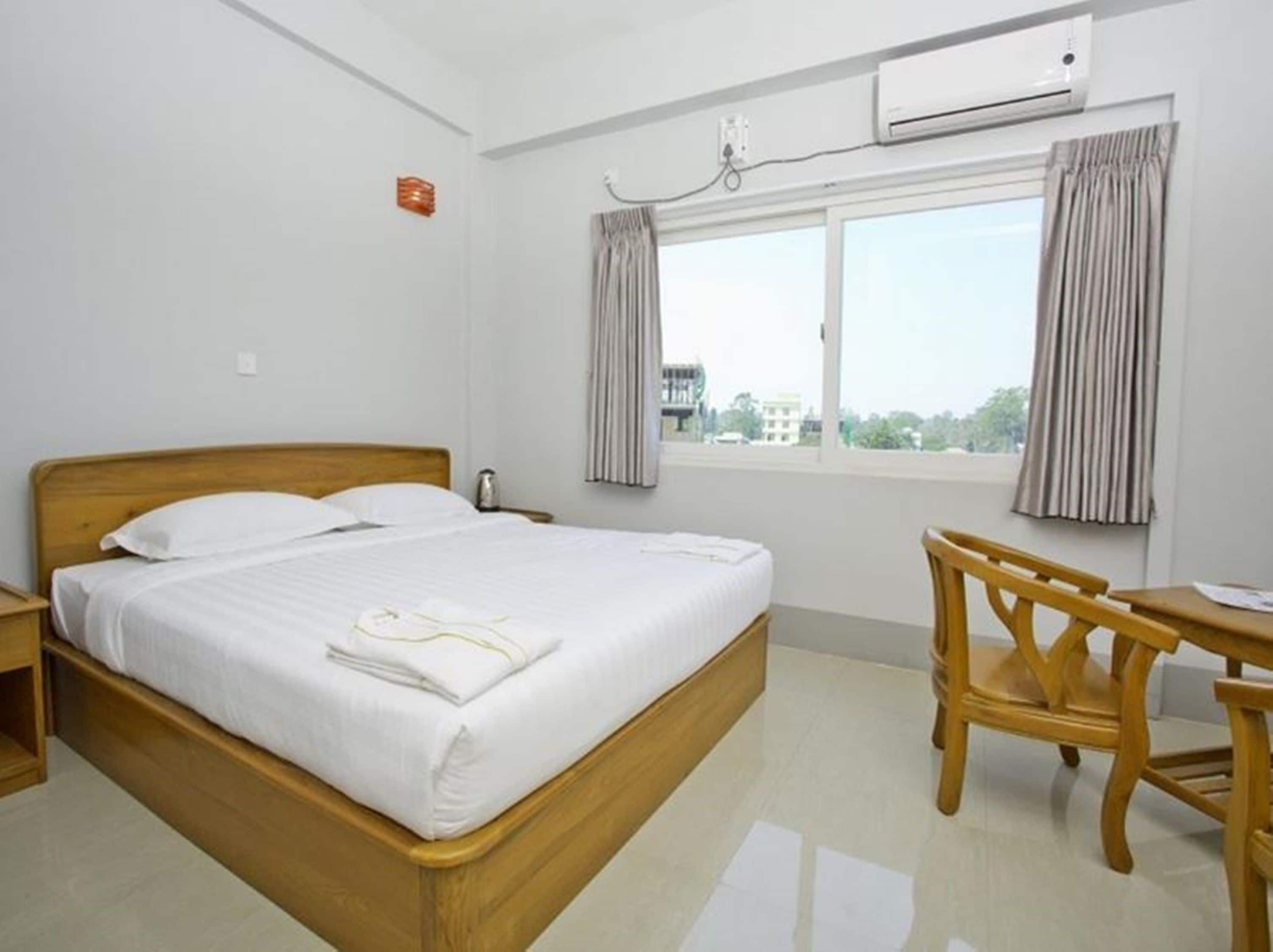 Habitación de hotel con cama de madera, un escritorio y una silla. Se observa una unidad de aire acondicionado en la pared y una ventana con cortinas.