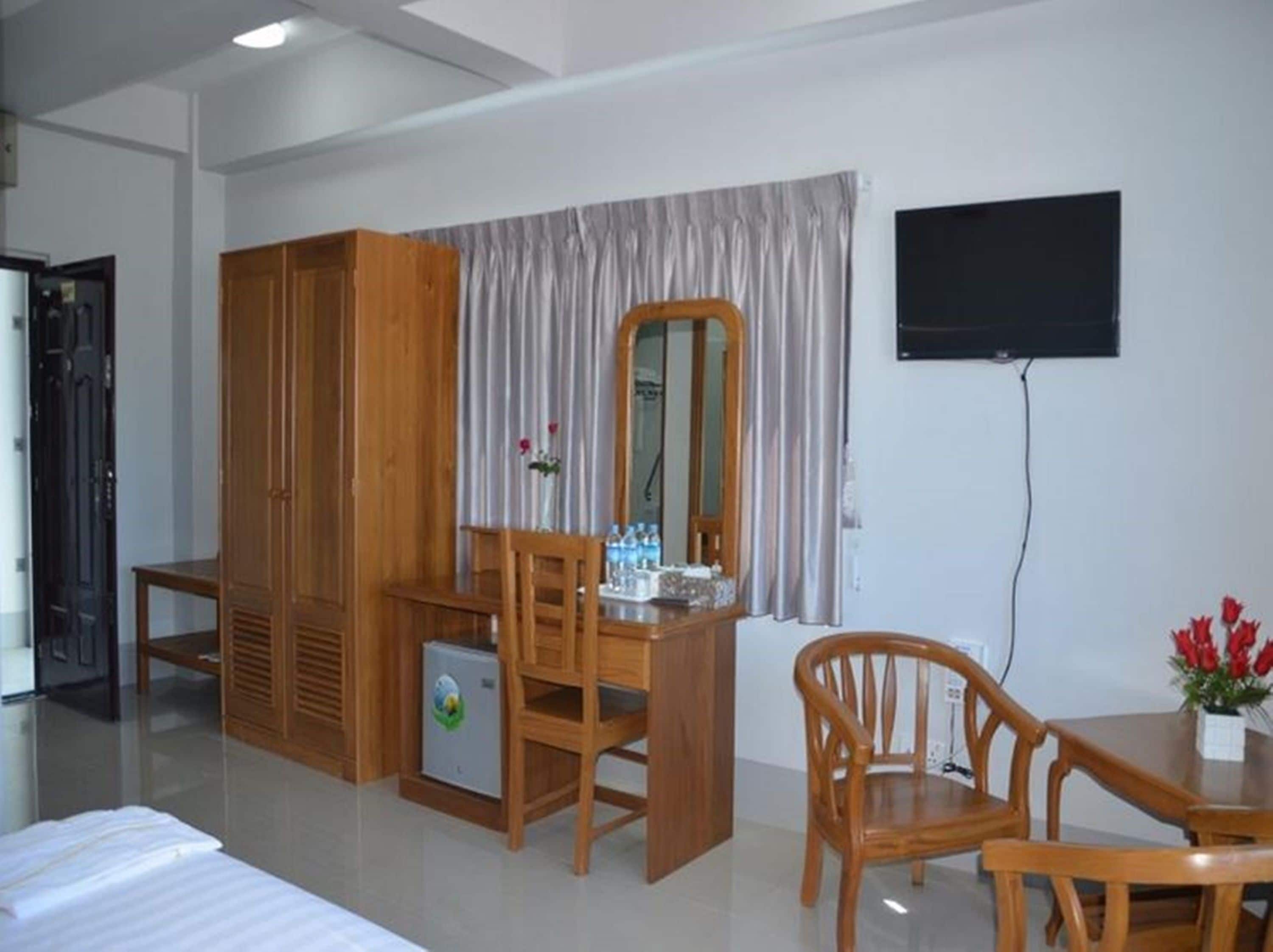 A hotel room with a bed, wooden wardrobe, desk, chairs, a small refrigerator, a mirror, a television, and a window with curtains.