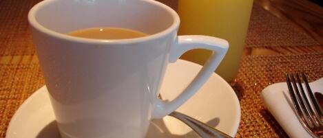 Una taza blanca de café sobre un plato con una cuchara, junto a un vaso de jugo de naranja sobre una mesa.