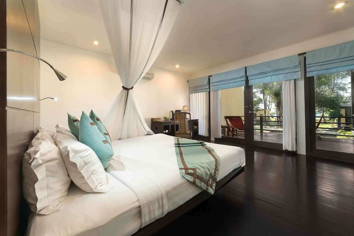 A bedroom with a bed, white curtains, a wooden floor, and a view of the outdoors.