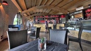 Een restaurantruimte met houten balken in het plafond, rieten stoelen en een bar met een toog en flessenplanken.