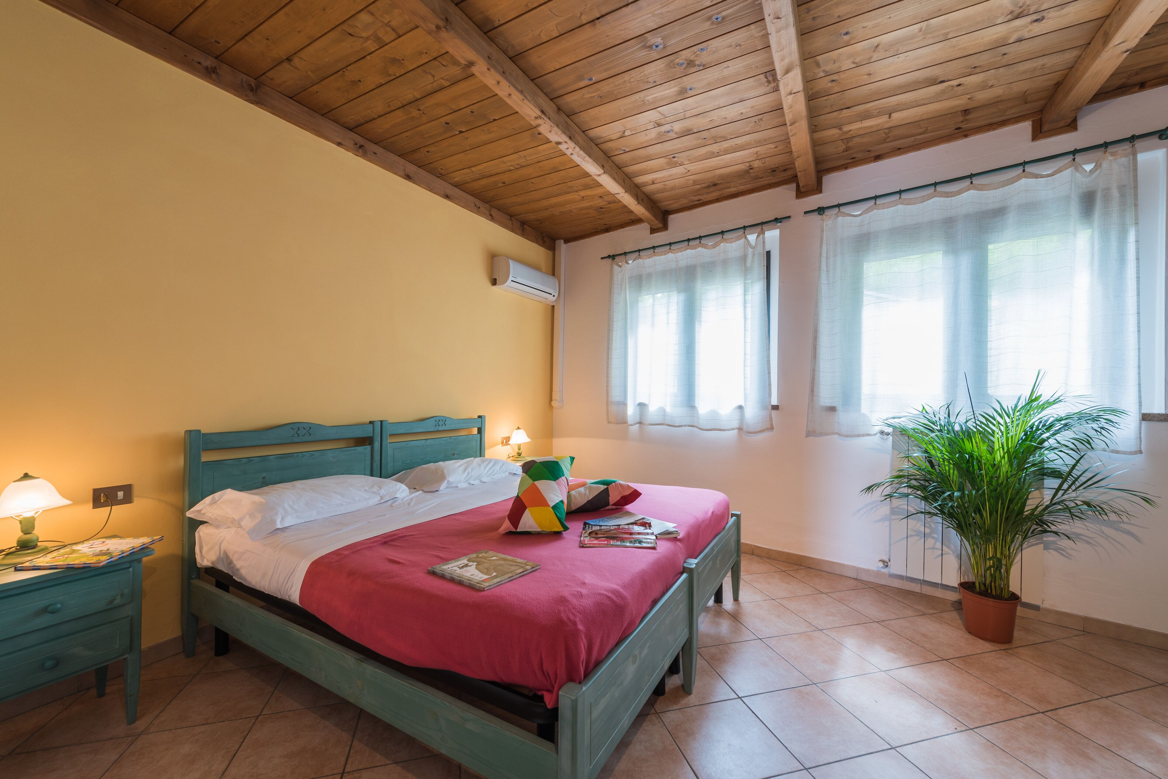 Un dormitorio con techo de madera, una cama con colcha roja, una mesita de noche con lámpara y una planta en maceta.