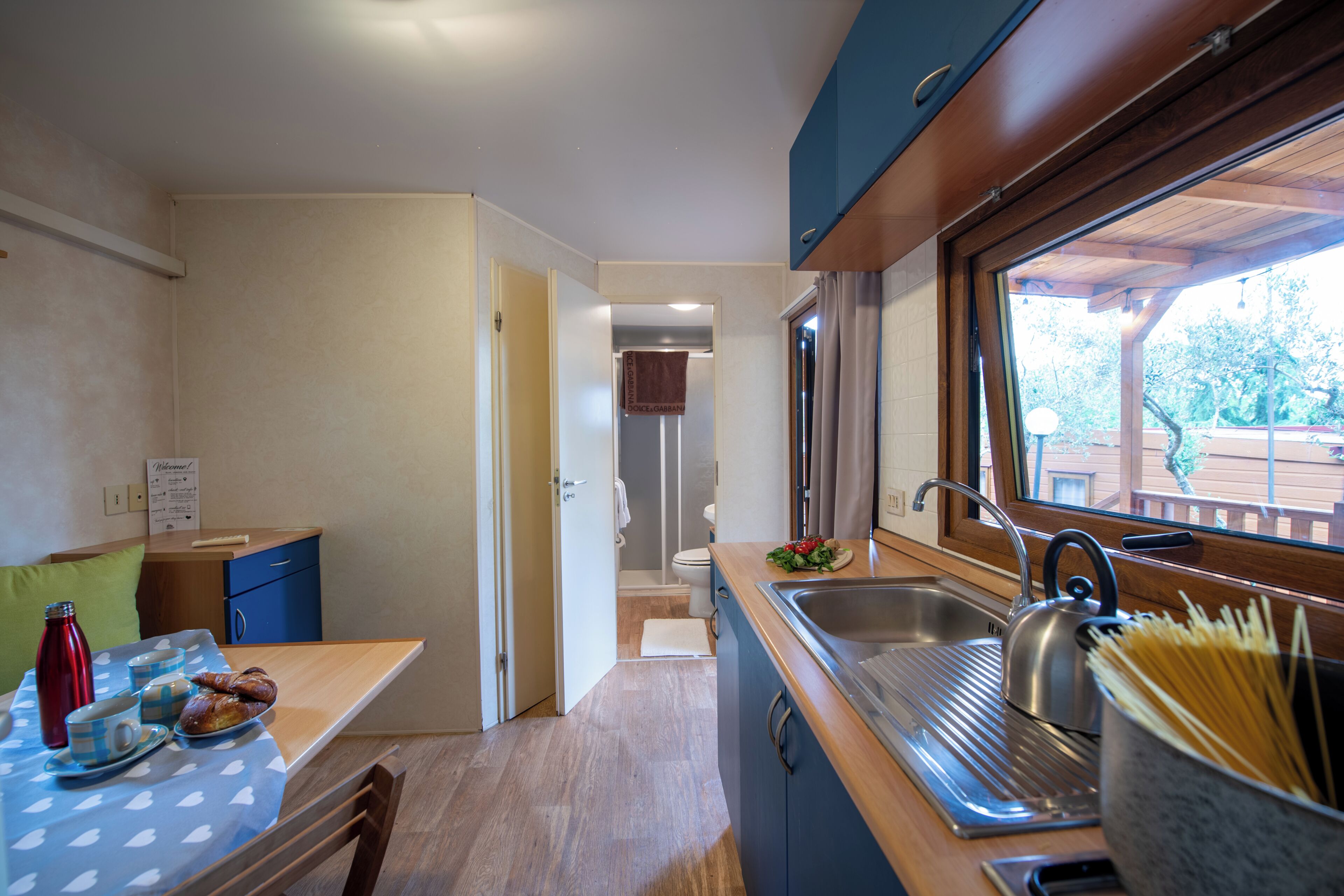 A compact kitchen with a sink, a window with a view, a table with breakfast items, and a door leading to a bathroom.
