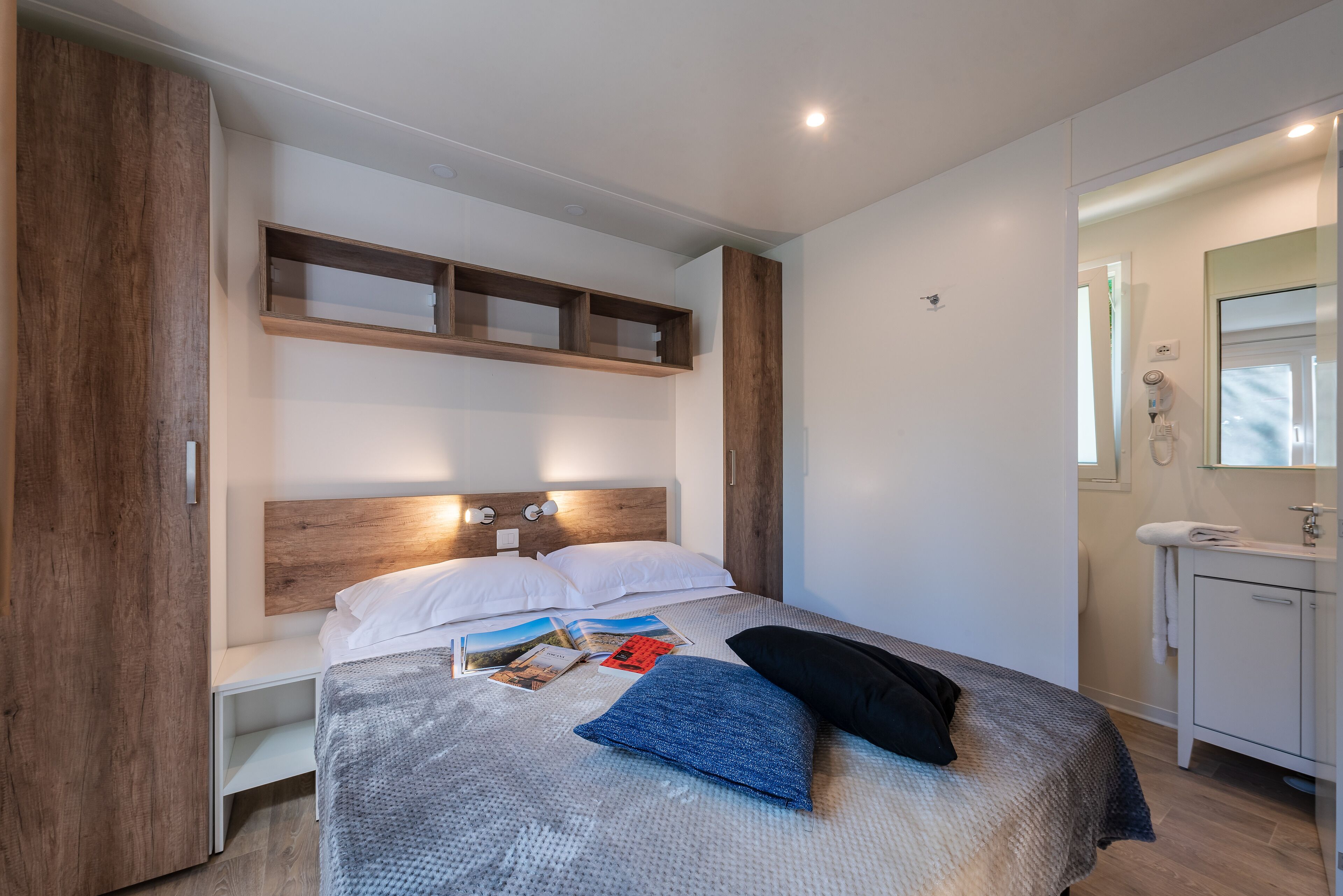 A modern bedroom with a wooden headboard, a bedside table, a built-in shelf, and a small bathroom with a mirror and sink.