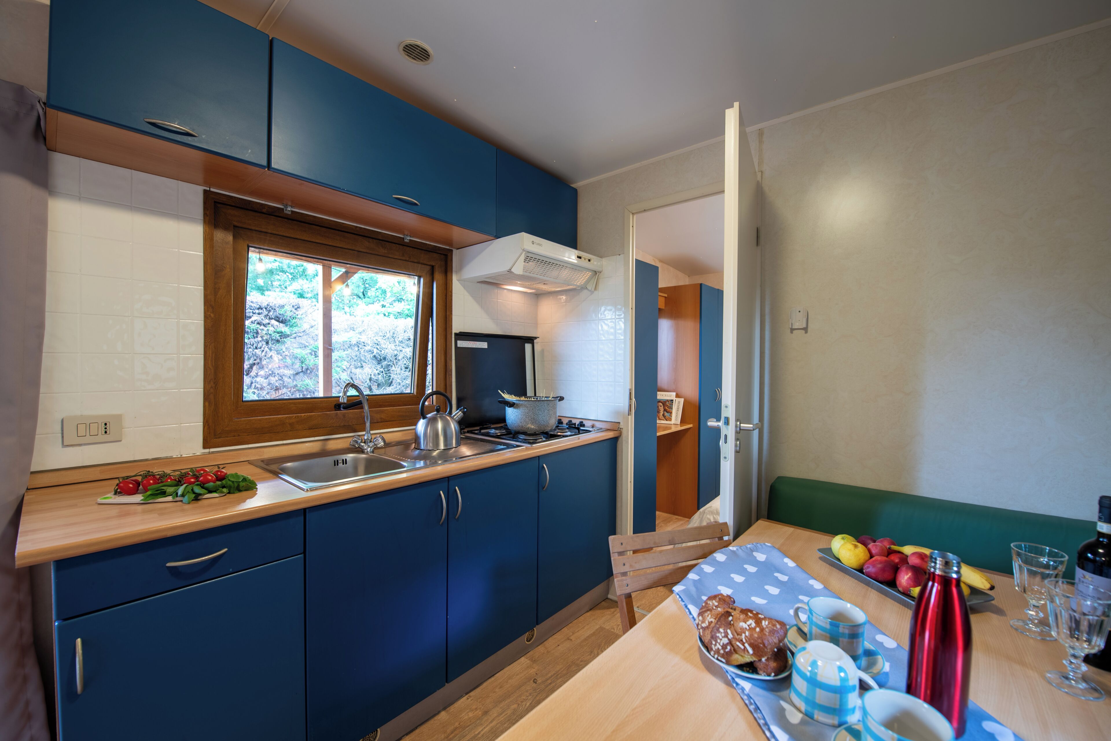 A kitchen with blue cabinets, a wooden countertop, a window, and a dining area with a table set for two.