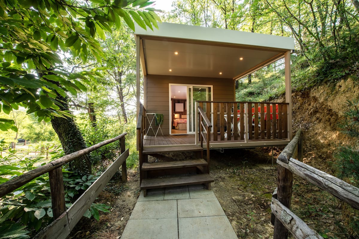 Une terrasse en bois mène à un chalet doté d’un porche abrité, le tout étant entouré d’une forêt dense.