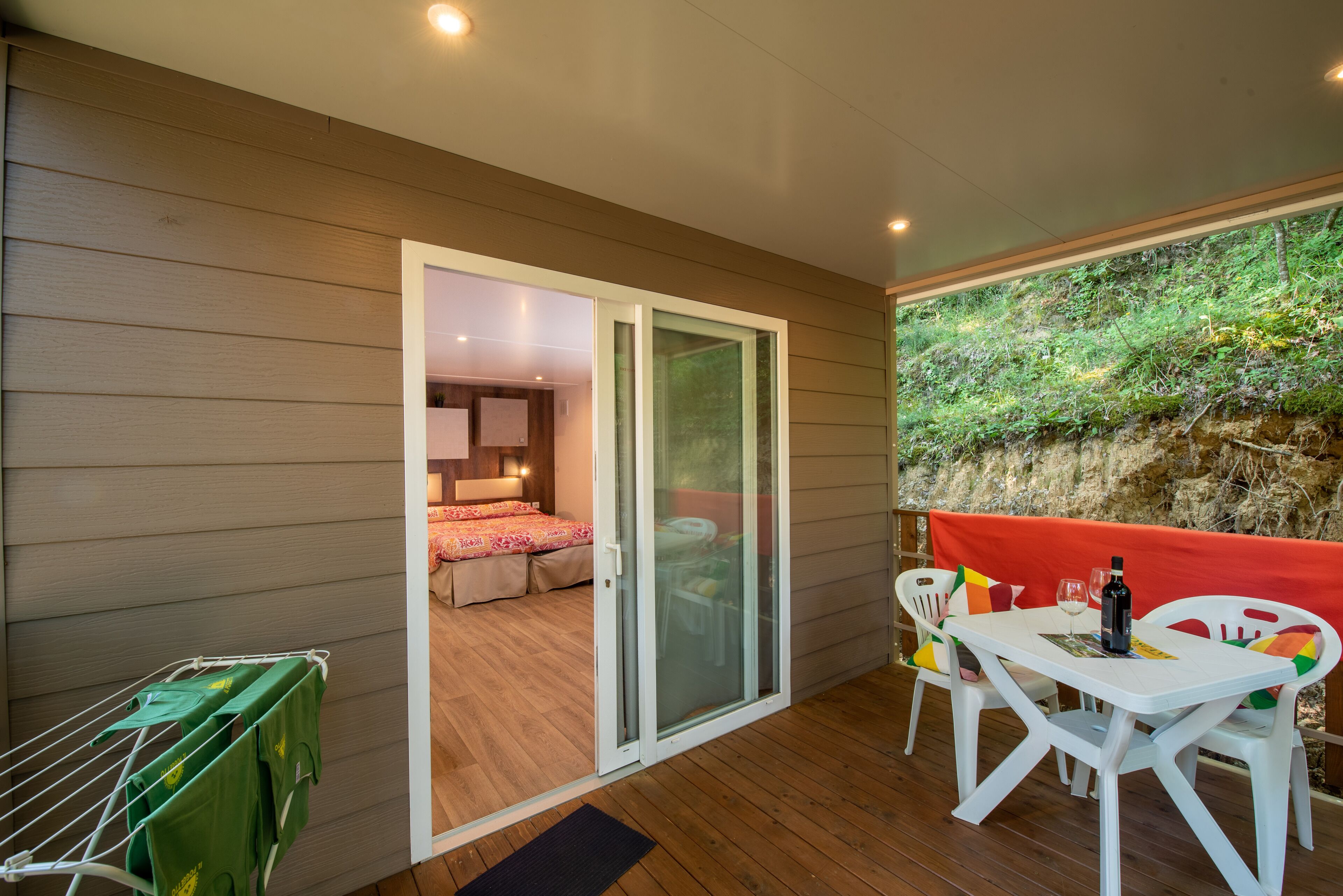 A room with a sliding glass door leading to a balcony with a table and chairs, a bed with a red headboard, and a drying rack with green towels.