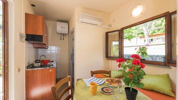 A small kitchen with a stove, sink, and wooden cabinets. There is a dining table with a green tablecloth, a potted plant, and a window with a view of the outside.