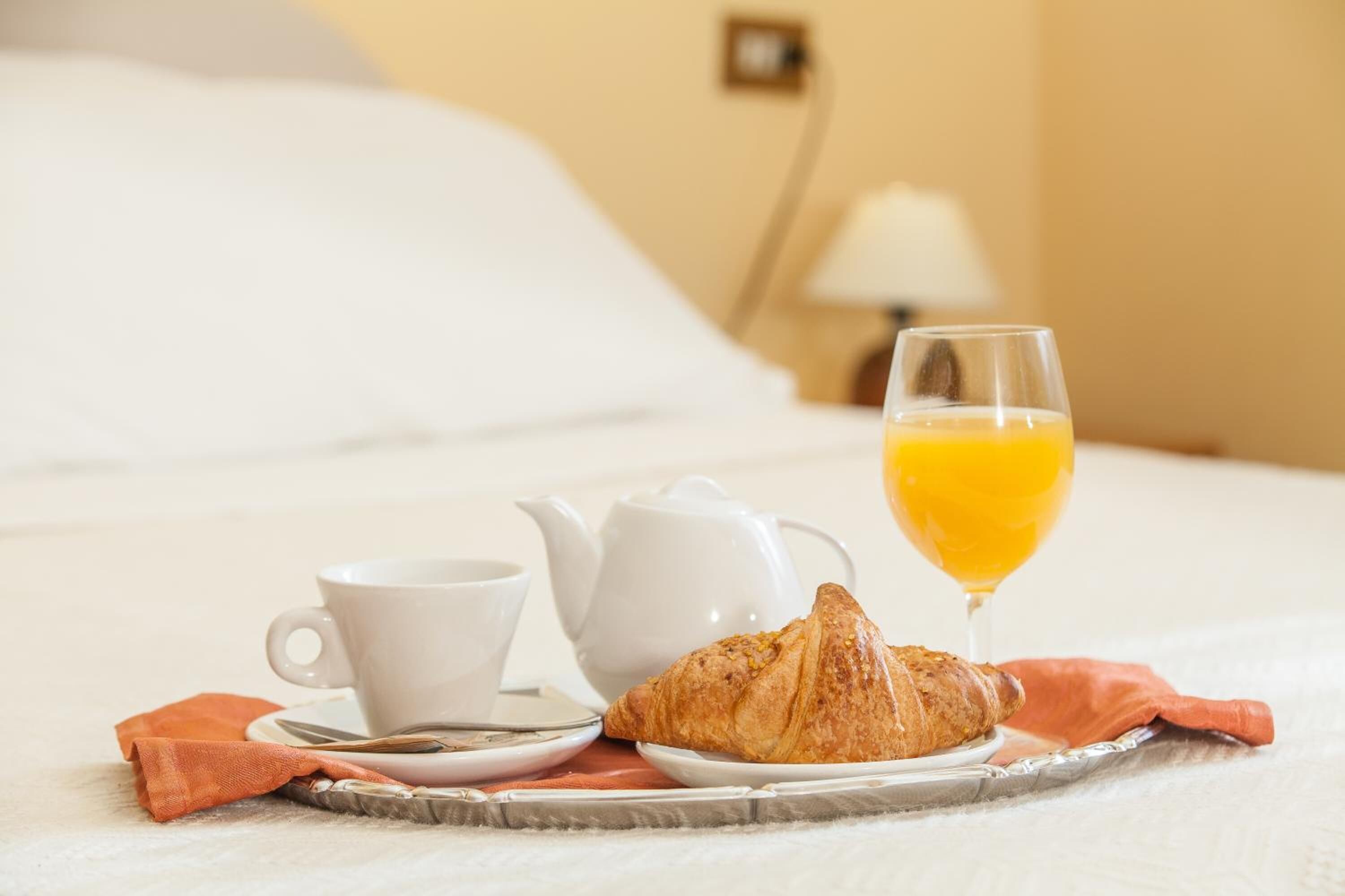 A breakfast setup on a bed with a croissant, a cup, a teapot, and a glass of orange juice.