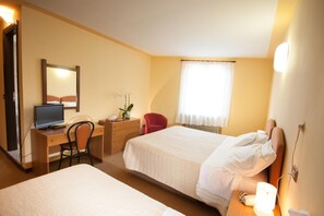 A hotel room with two beds, a desk with a computer, a chair, a small table with a plant, and a window with curtains.