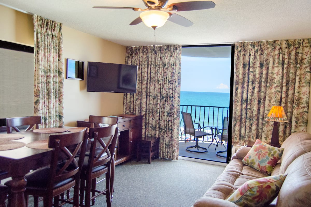 A hotel room with a dining area, a sofa, a TV, and a balcony with a view of the ocean.
