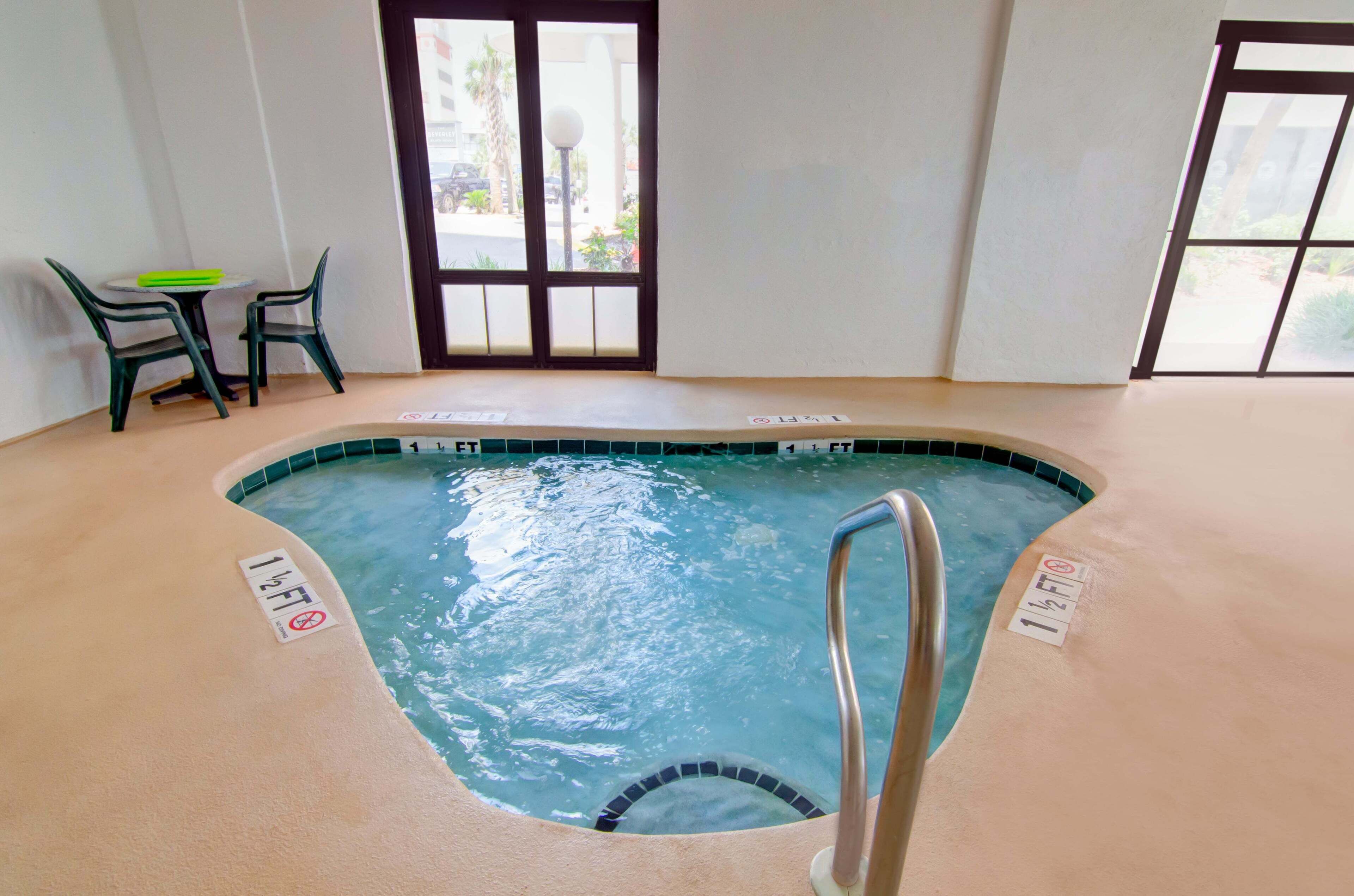 Ein Hallenbad mit einem Hinweisschild für eine Wassertiefe von 1,5 Metern, einem kleinen Tisch mit Stühlen und einer Tür mit Fenstern.