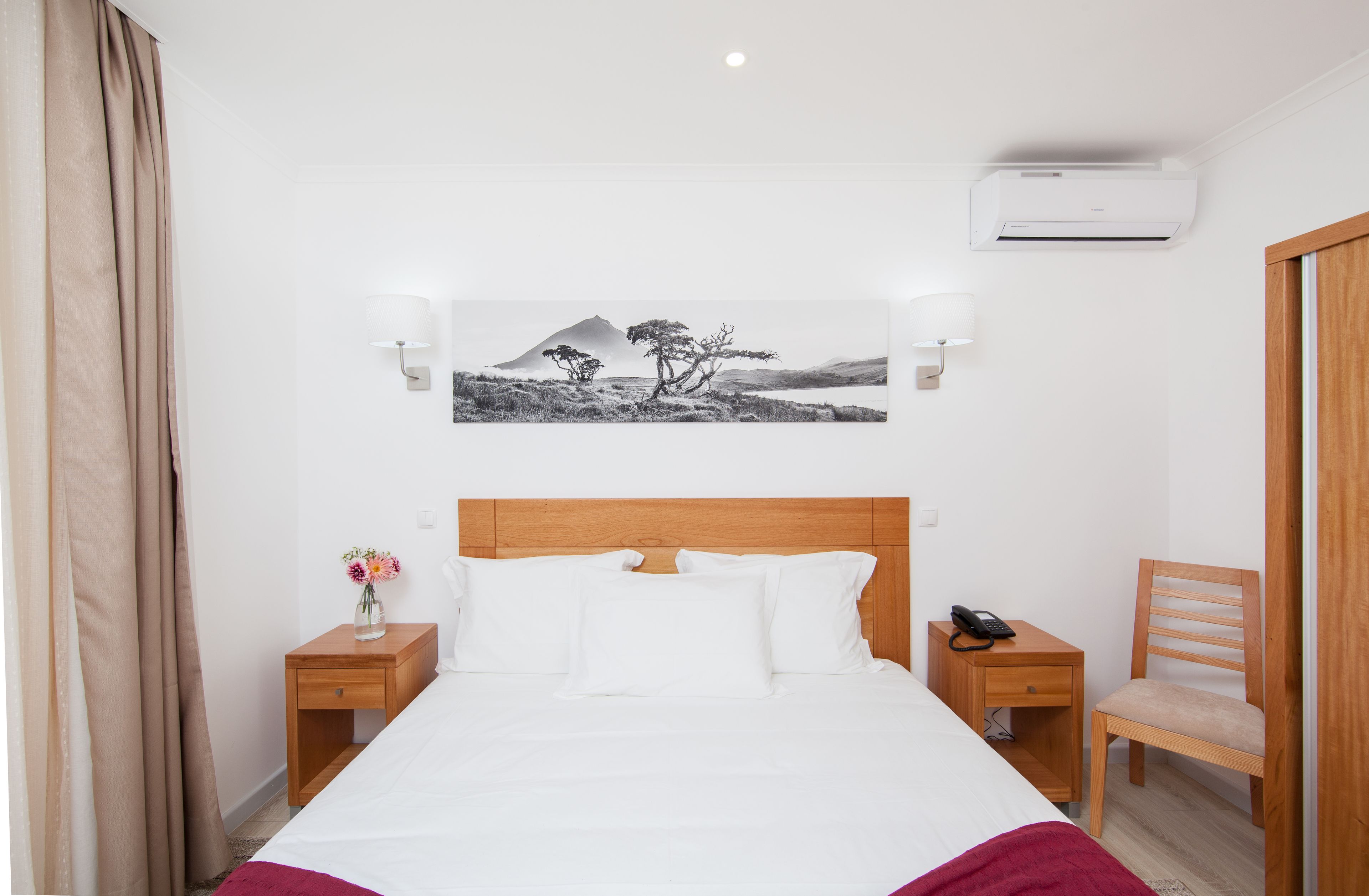 A hotel room with a bed, two bedside tables, a nightstand, a chair, a wall-mounted air conditioner, and a framed picture on the wall.