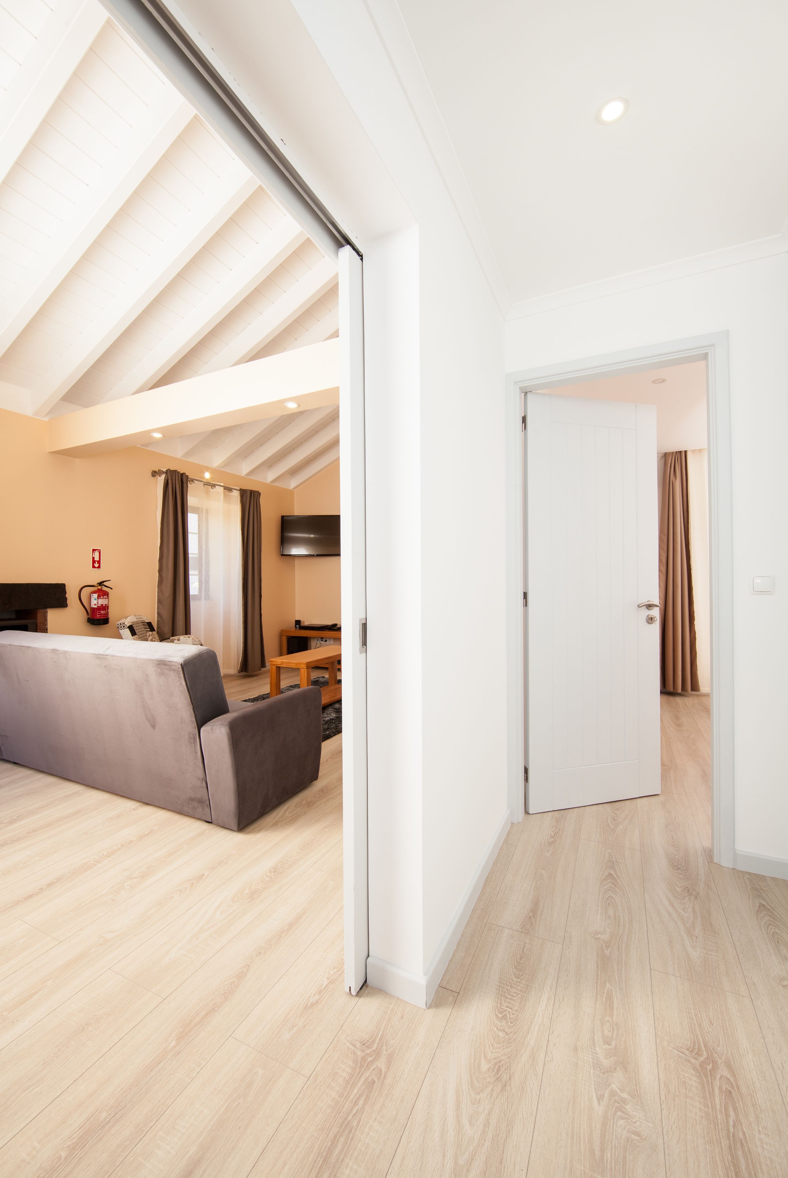 Quarto moderno com piso de madeira, sofá, televisão e porta para outro espaço.