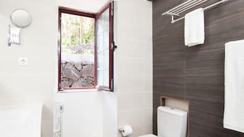 A modern bathroom with a toilet, bidet, sink, and a window with a stone wall view.