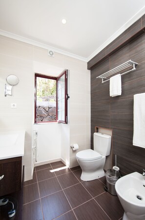 A modern bathroom with a toilet, bidet, sink, and a window with a stone wall view. - Aldeia das Adegas (Sao Roque do Pico)