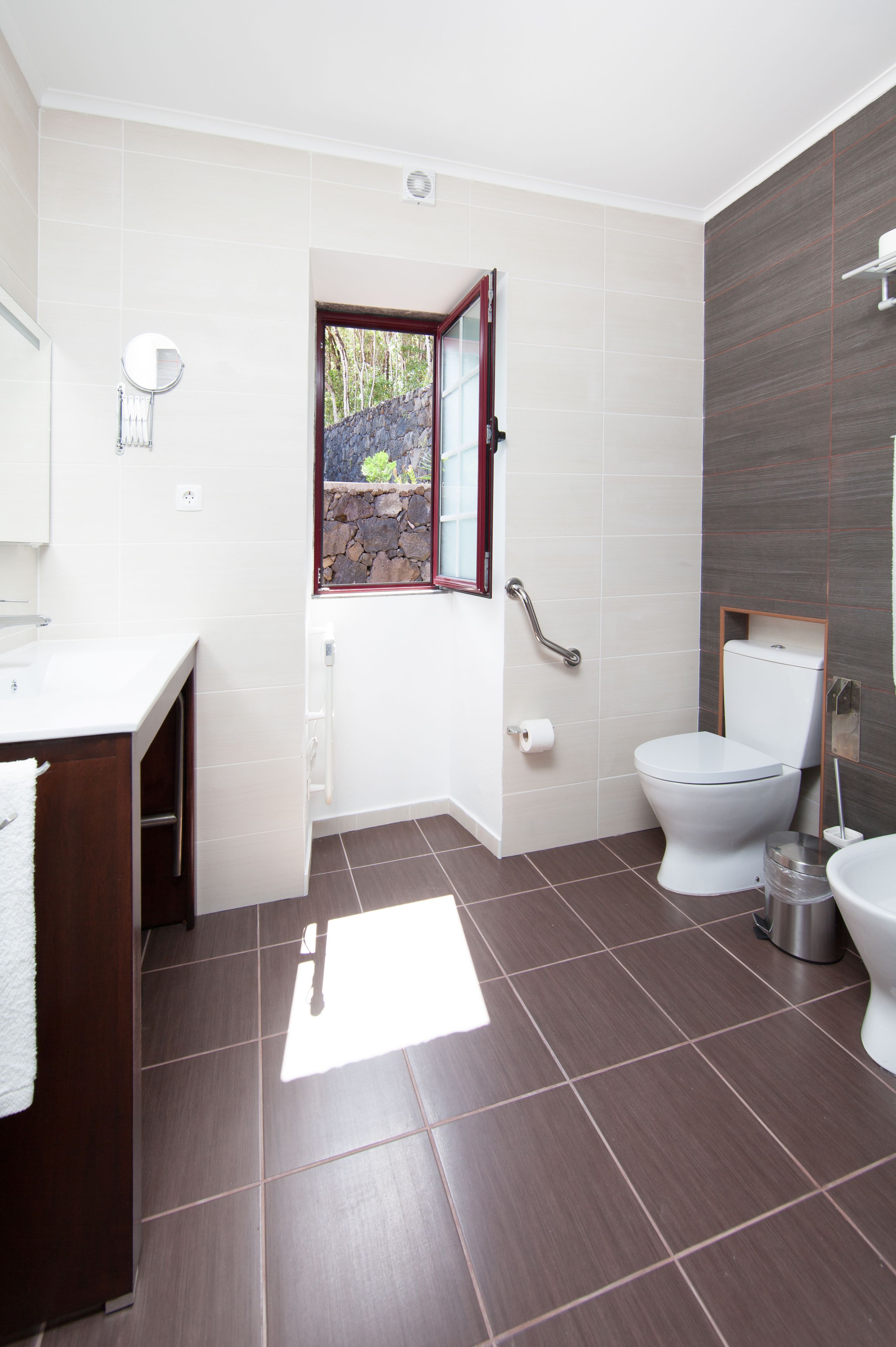 Une salle de bain moderne avec des toilettes blanches, un bidet, un lavabo avec un miroir et une fenêtre donnant sur un paysage verdoyant.