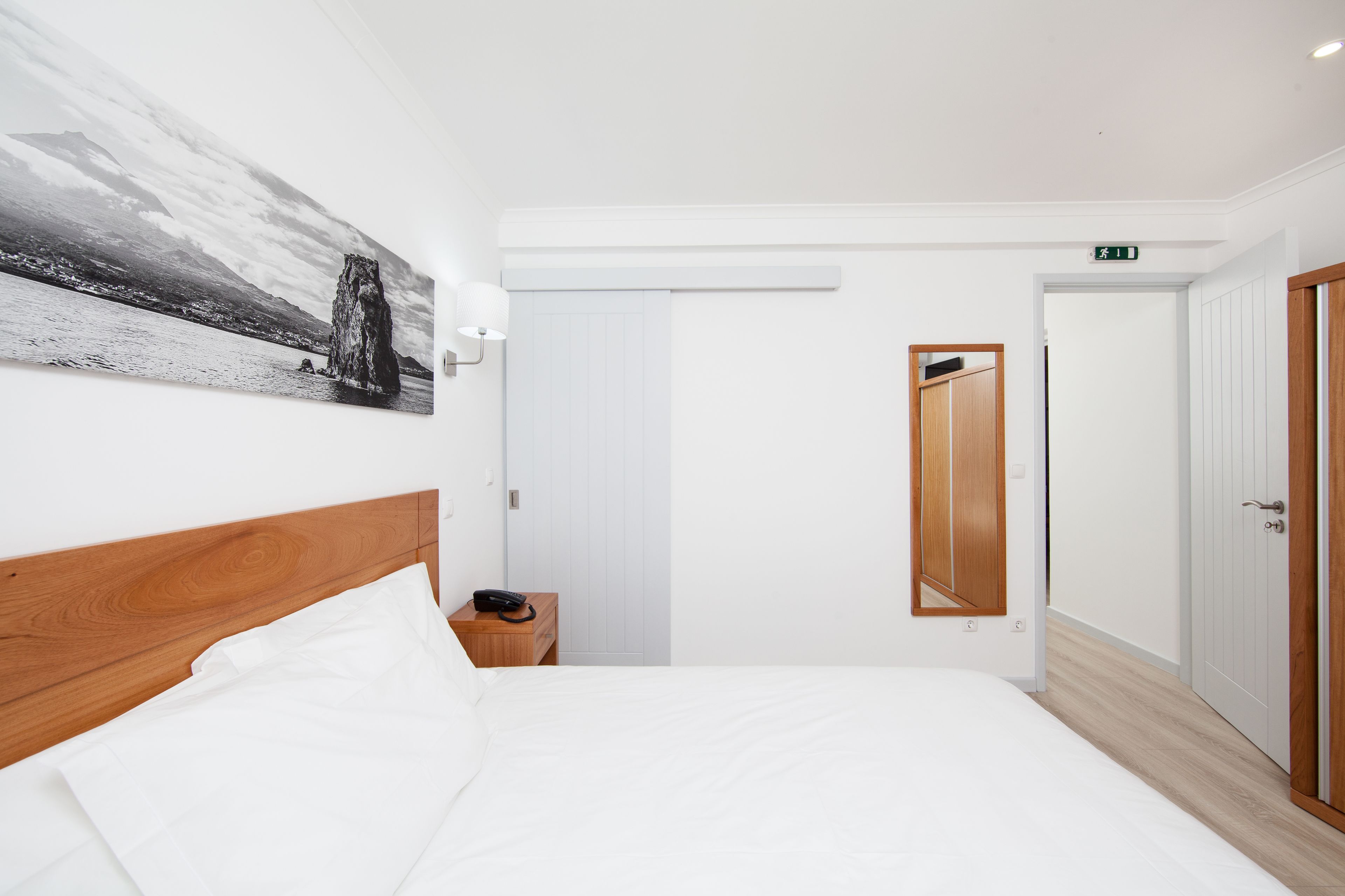 Habitación de hotel con cabecera de madera, cama blanca, mesita de noche con teléfono, pintura de un paisaje, puerta blanca y espejo.