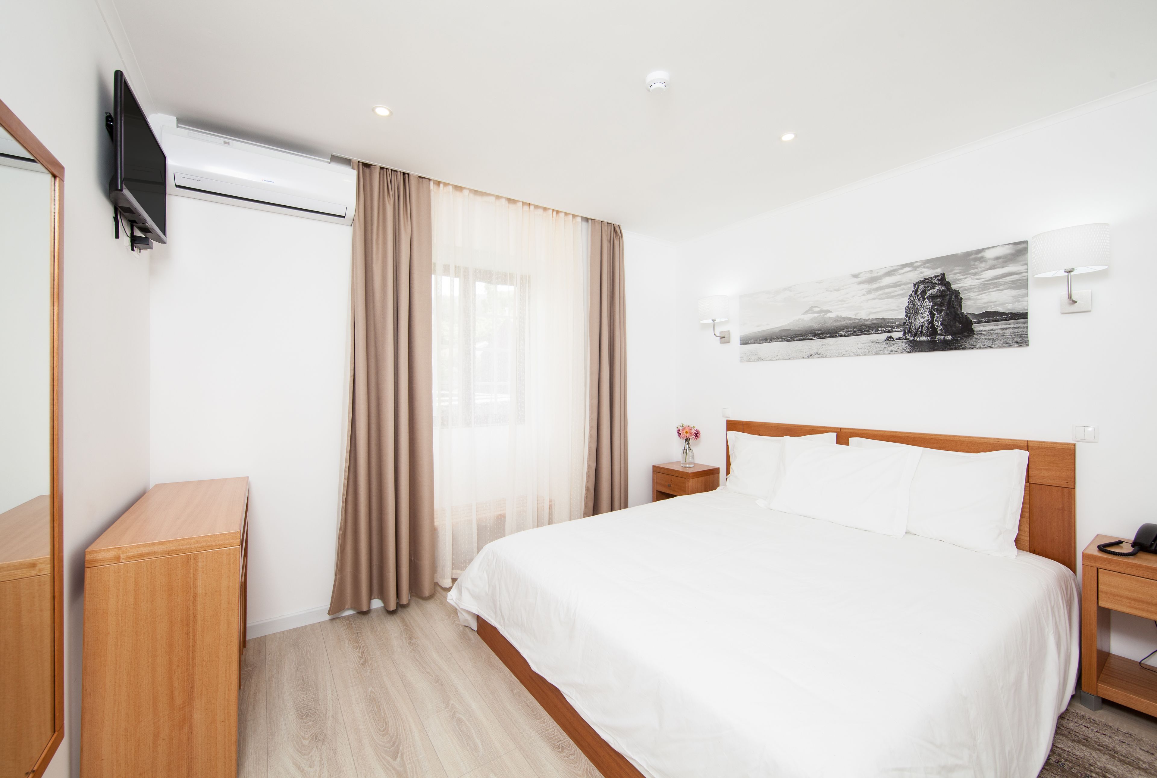 A hotel room with a bed, a flat-screen TV mounted on the wall, a bedside table with a lamp, and a painting on the wall.