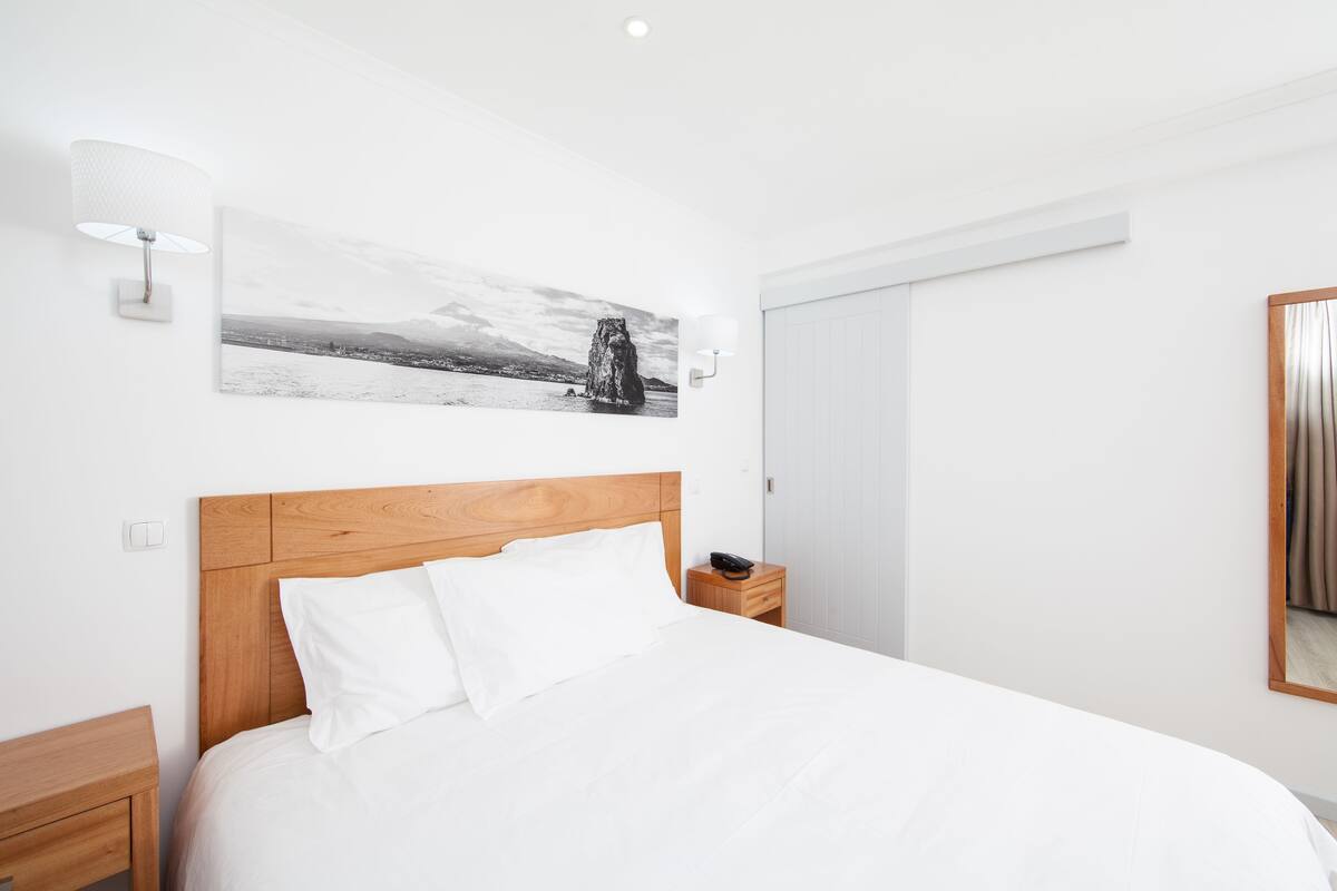Un dormitorio moderno con cabecera de madera, ropa de cama blanca, una mesita de noche con lámpara y una fotografía enmarcada en la pared.