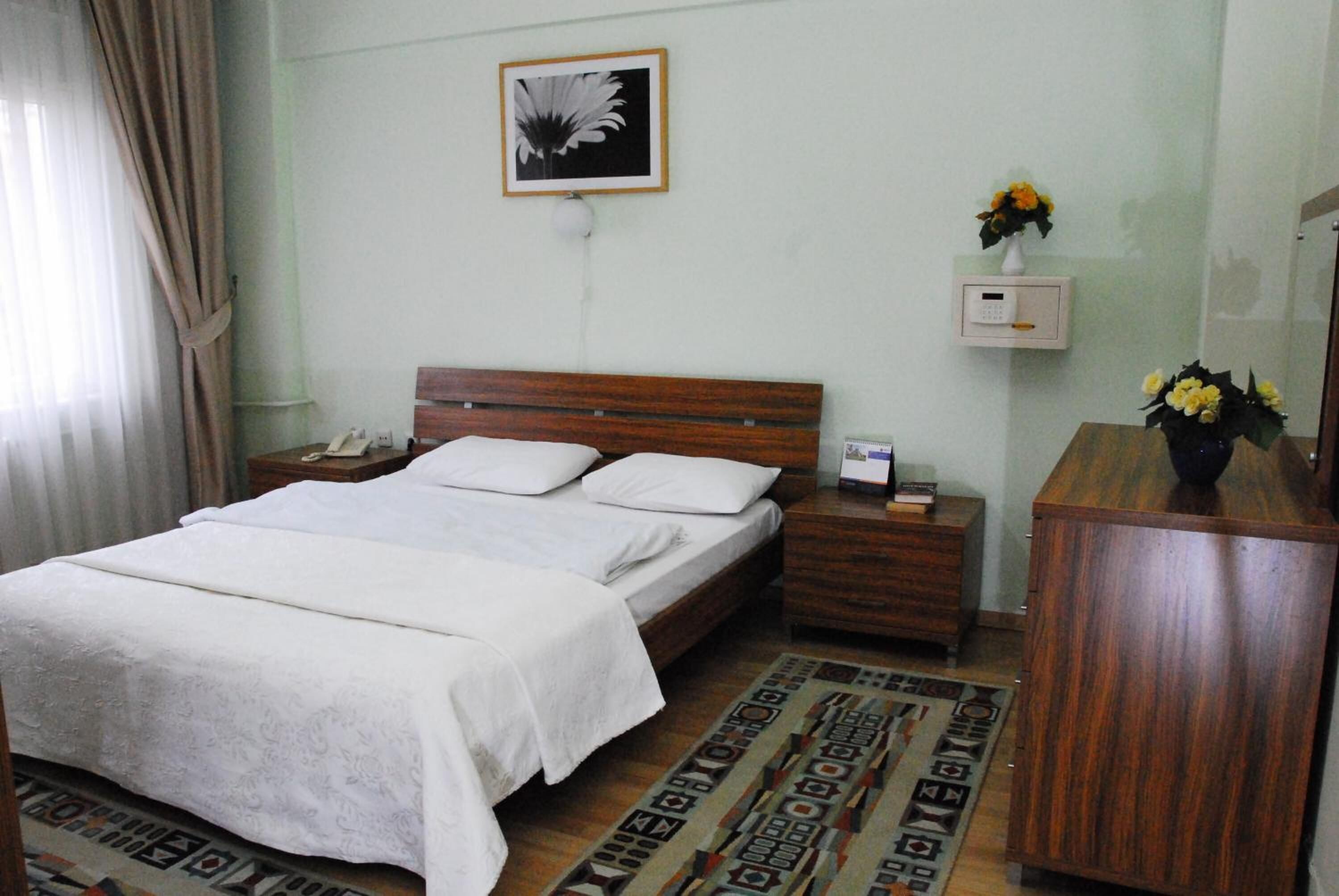Habitación de hotel con cama, mesitas de noche, termostato en la pared, un cuadro enmarco y un florero con flores.