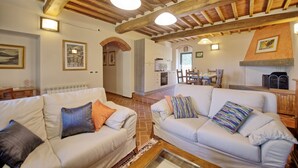 Ein geräumiges Wohnzimmer mit zwei weißen Sofas, einem Essbereich und einer Küche mit freiliegenden Holzbalken und Backsteinwänden.