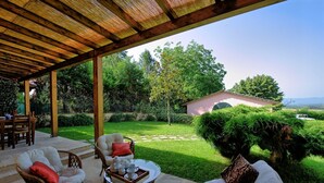 Eine überdachte Terrasse mit Gartenmöbeln, einem Esstisch und Blick auf einen Garten und ein Haus im Hintergrund.