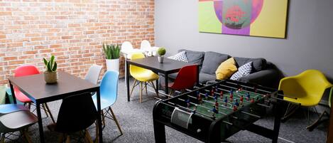 A room with a brick wall, a sofa, colorful chairs, a foosball table, and a table with a plant.