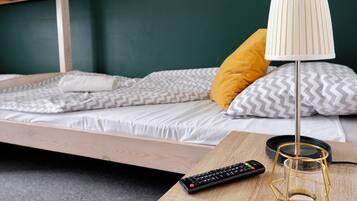 Un lit simple avec un cadre en bois, un oreiller blanc, un oreiller jaune, une lampe blanche et une télécommande posée sur une table de chevet en bois.