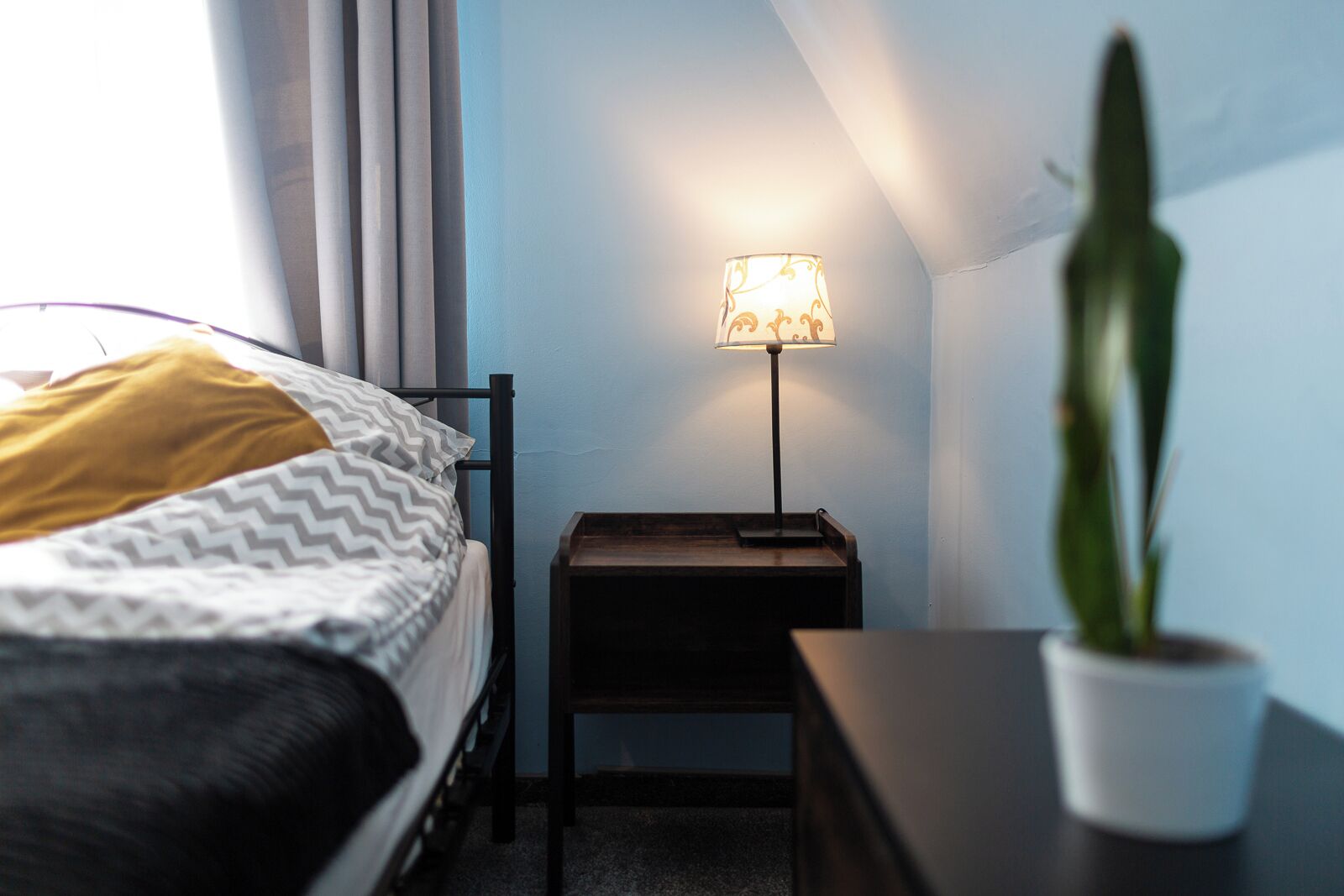 Un dormitorio con una cama, una mesita de noche con lámpara, una planta en maceta y un ventanal con cortinas.