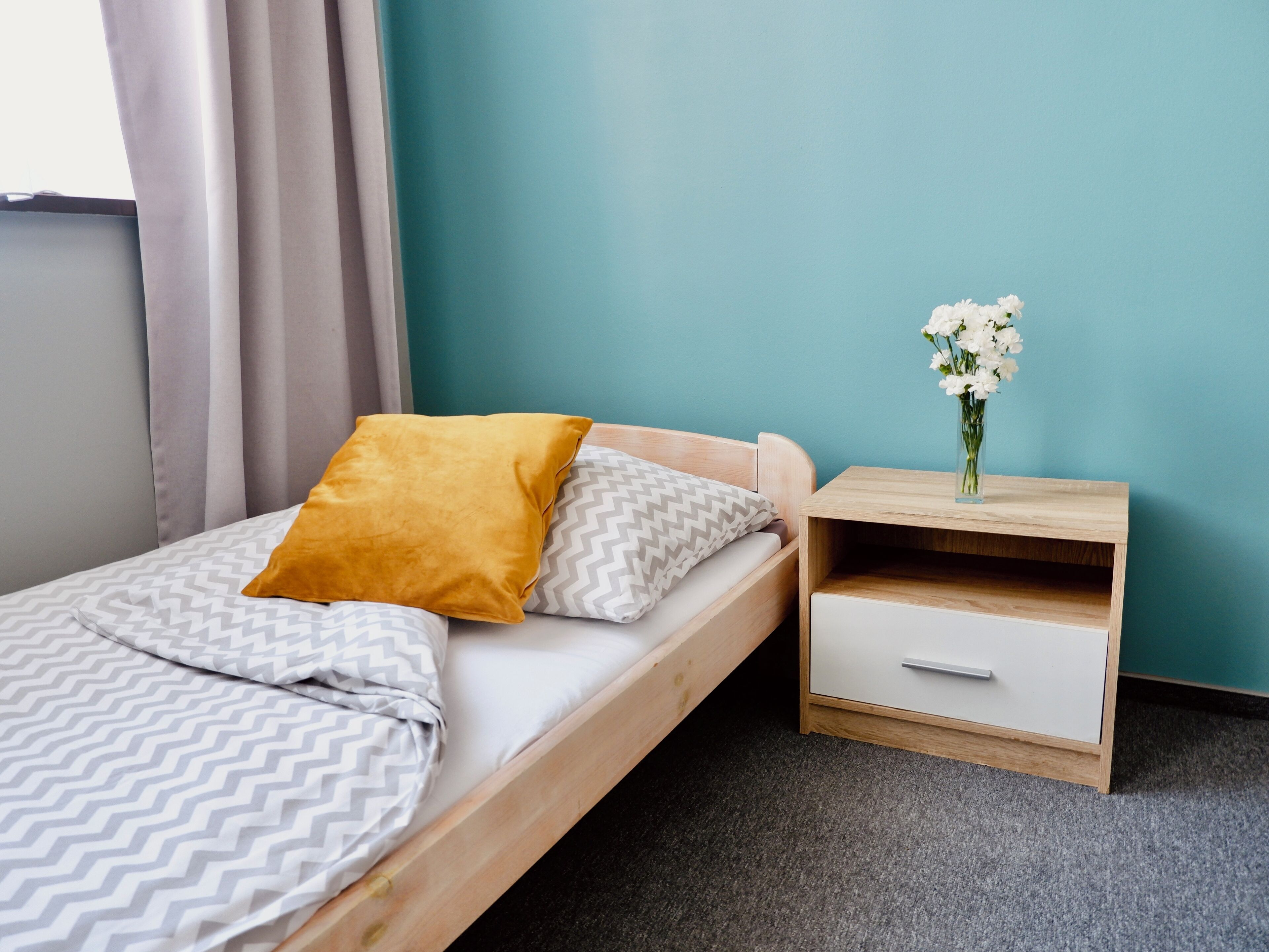 Ein Einzelbett mit Holzrahmen, eine Decke in Weiß und Grau gemustert, ein senfkörnermelangefarbenes Kissen und ein weiteres Kissen in Weiß und Grau gemustert. Ein Nachttisch aus Holz mit einer Schublade und eine Vase mit weißen Blumen darauf. Die Wand ist teal gefärbt, der Boden ist mit Teppich ausgelegt.