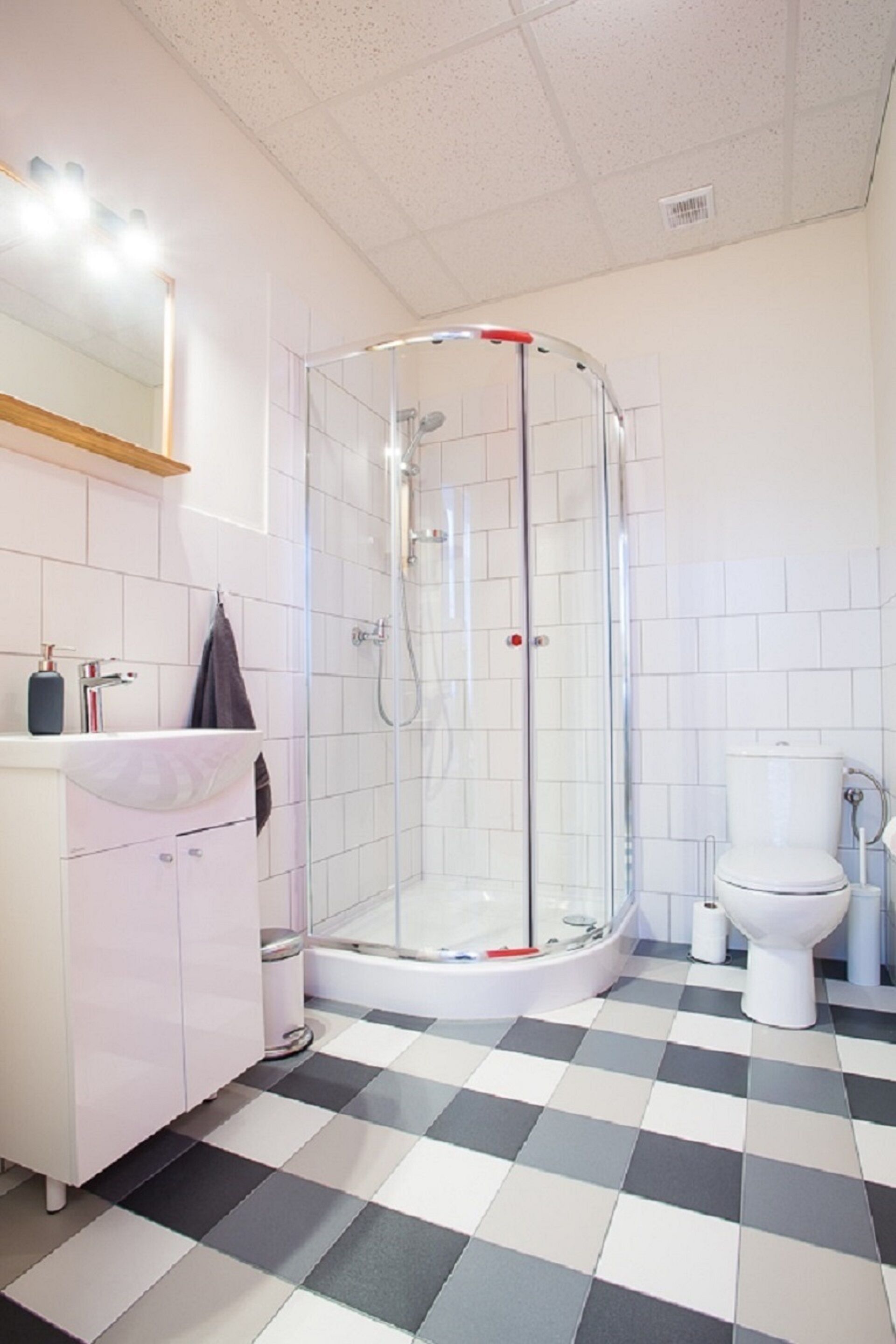Ein Badezimmer mit einer gläsernen Duschkabine, einem weißen Waschbecken mit Spiegel darüber und einer Toilette. Der Boden weist ein Schachbrettmuster auf.