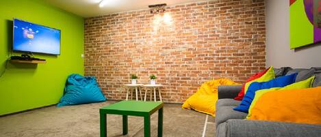 A room with a brick wall, a green table, a grey sofa with colorful cushions, a blue bean bag, and a TV on the wall.