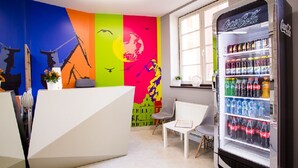 A brightly colored reception area with a Coca-Cola refrigerator stocked with beverages, a white reception desk, and a mural on the wall. - Moon Hostel Gdańsk (Gdansk)