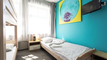A bedroom with a bunk bed, a single bed, a bedside table with a lamp, a TV mounted on the wall, and a window with curtains.
