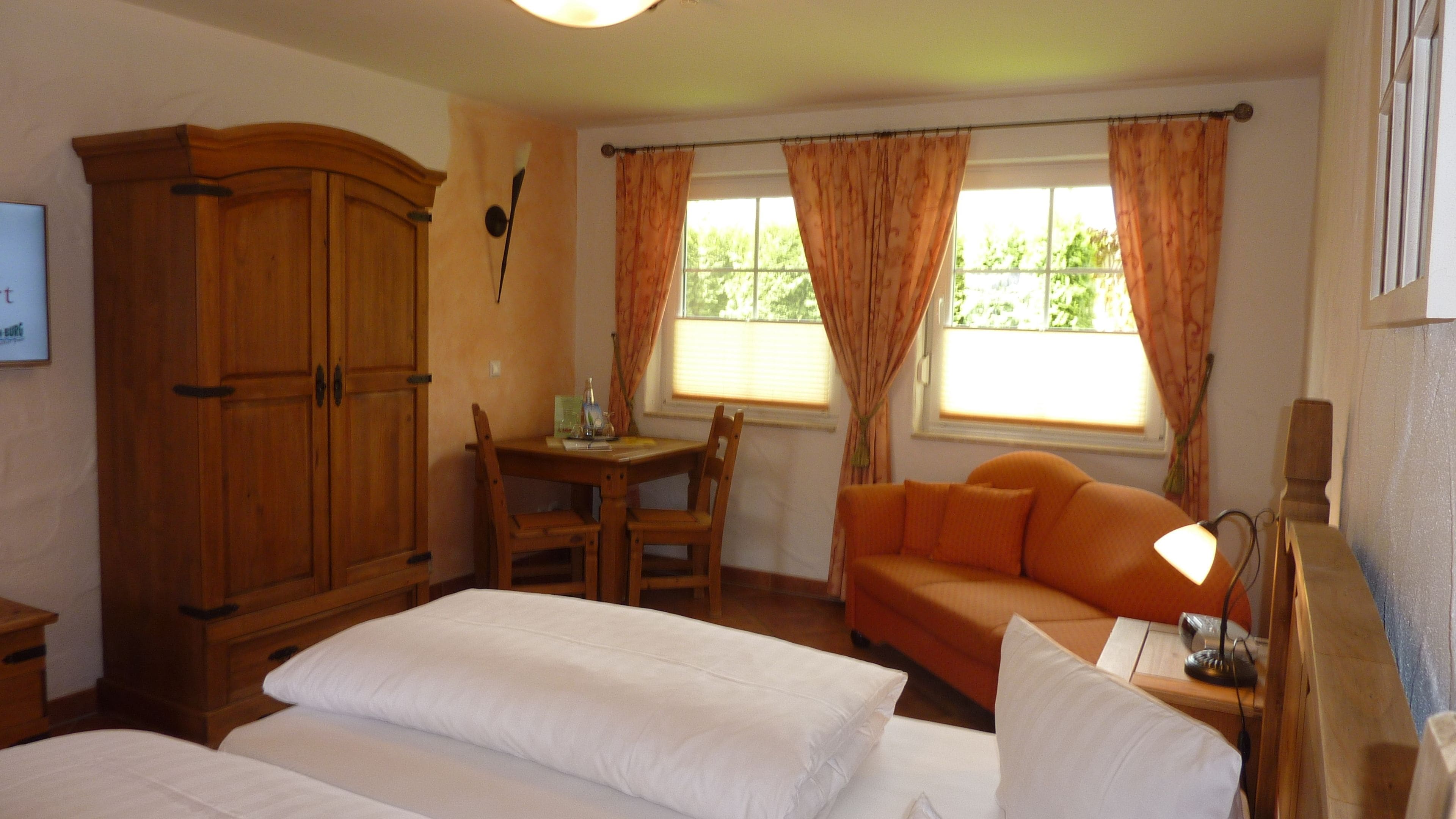A bedroom with two beds, a wooden wardrobe, a small table with chairs, an orange sofa, and a window with curtains.