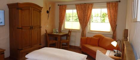 A bedroom with two beds, a wooden wardrobe, a small table with chairs, an orange sofa, and a window with curtains.