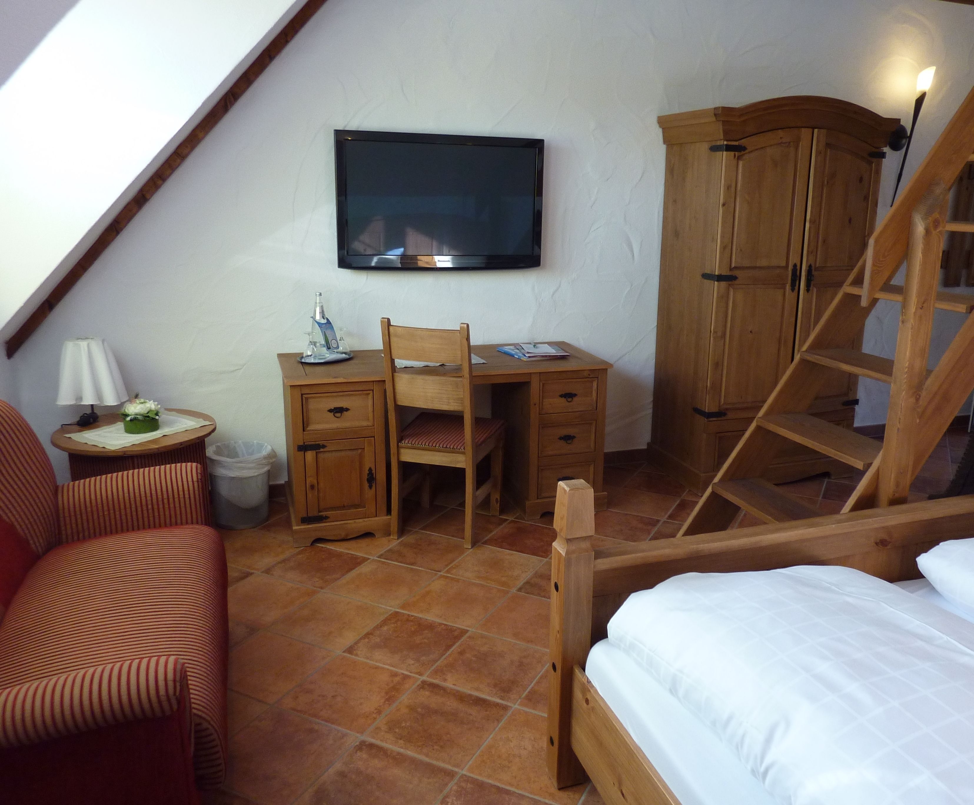 A bedroom with a wooden bed, a desk with a chair, a television on the wall, and a staircase.