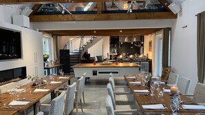 Un espace de repas moderne avec une longue table en bois dressée pour un repas, une cuisine en arrière-plan et un escalier menant à un niveau supérieur.