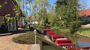 Un restaurant situé au bord de la rivière, avec une terrasse donnant sur l’eau, une enseigne publicitaire pour une pension, et une passerelle en bois menant aux tables.