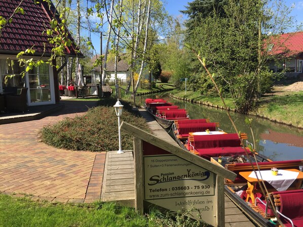 A riverside restaurant with outdoor seating along the water, a signboard advertising a pension, and a wooden walkway leading to the tables.